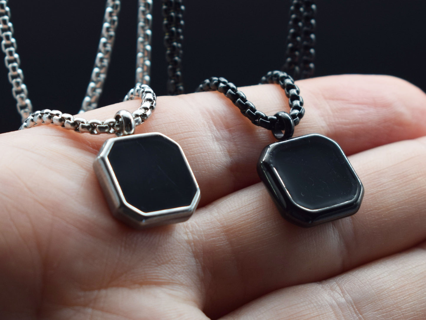 Two black silver pendant necklaces on a hand against a dark background
